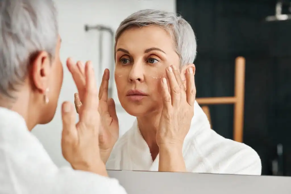 Woman applying moisturizer in the mirror at home.